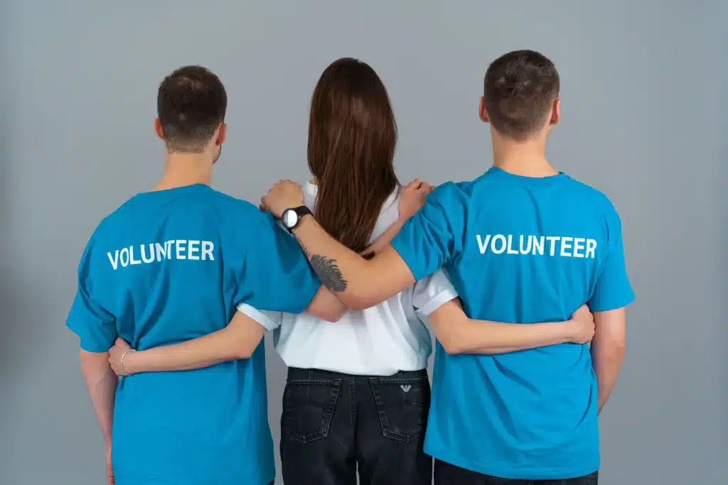 Photo de trois personnes alignées et de dos. Les personnes aux extrémités  portent un tee-shirt bleu arborant la mention “volunteer”, et la femme au centre porte un tee-shirt blanc. 