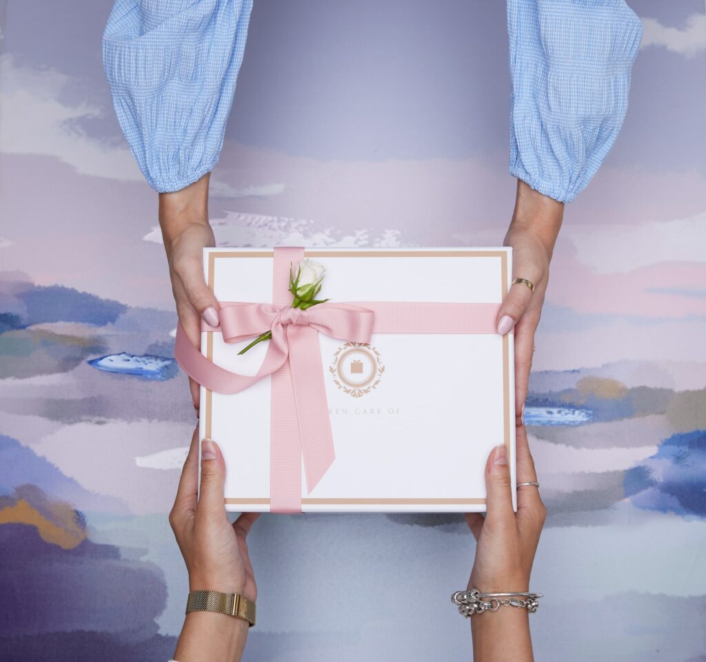 Boîte cadeau avec un ruban rose et une rose blanche passant de main en main avec un fond de couleurs pastel. 