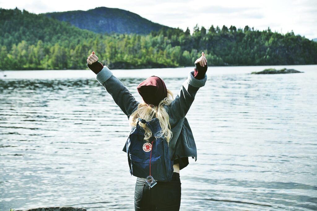Femme levant les bras en l'air avec un sac à dos devant un lac avec en arrière plan une forêt et une montagne.